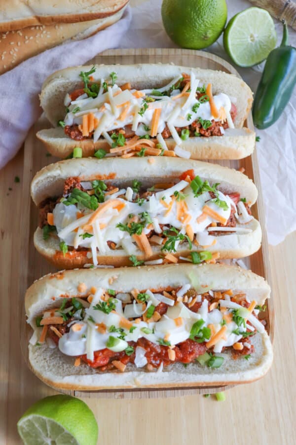 Hot dogs with taco toppings on a wooden cutting board.