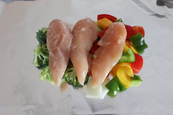 Three pieces of raw chicken laying on top of raw vegetables on aluminum foil.