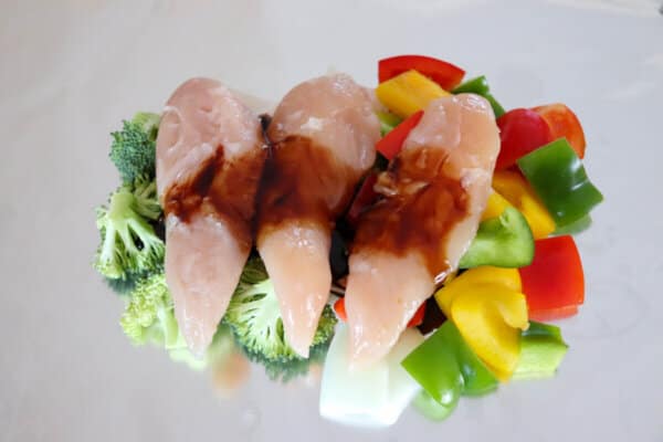 Three pieces of raw chicken laying on top of raw vegetables  with teriyaki sauce on aluminum foil.