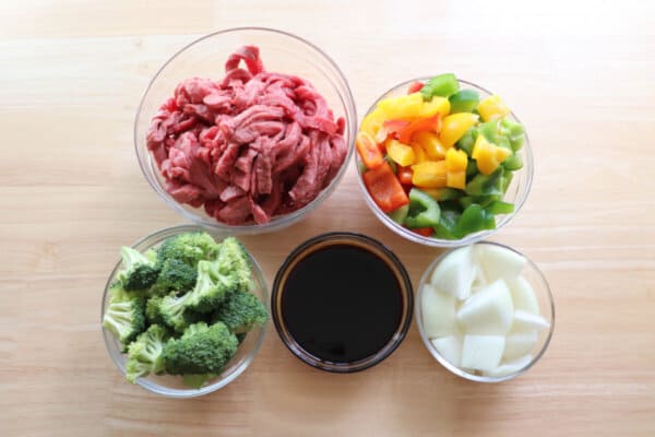 Ingredients for teriyaki steak foil packet meal in clear bowls on a wood surface.