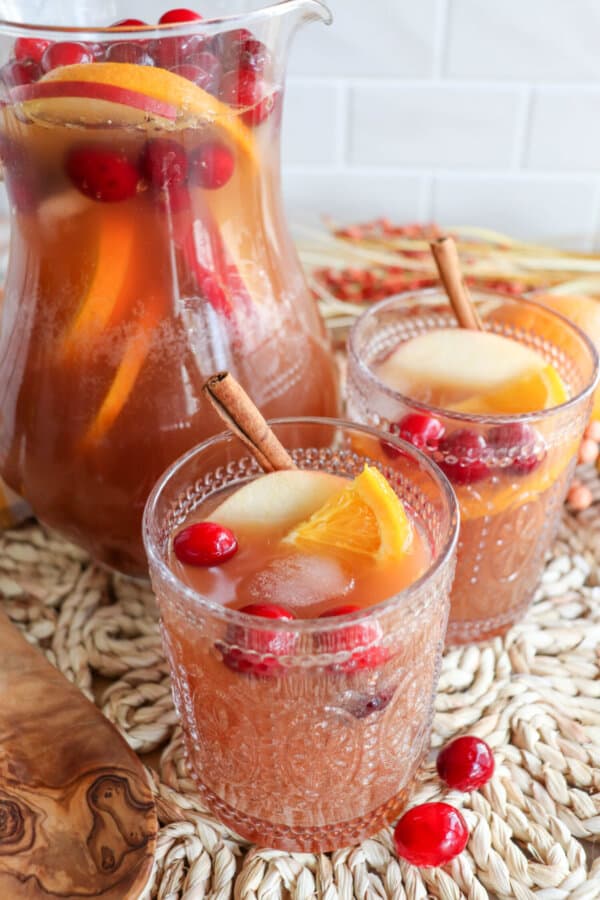 Thanksgiving punch in a glass pitcher with two filled glasses topped with apple slices, orange slices and cranberries.