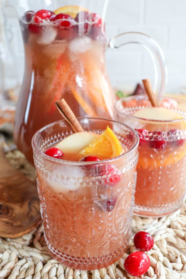 Thanksgiving punch in a glass pitcher with two filled glasses topped with apple slices, orange slices and cranberries.