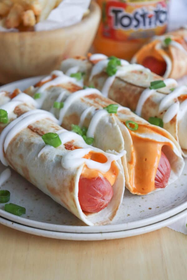 A close up of three hot dogs wrapped in tortillas on a cream plate garnished with crema and green onions.