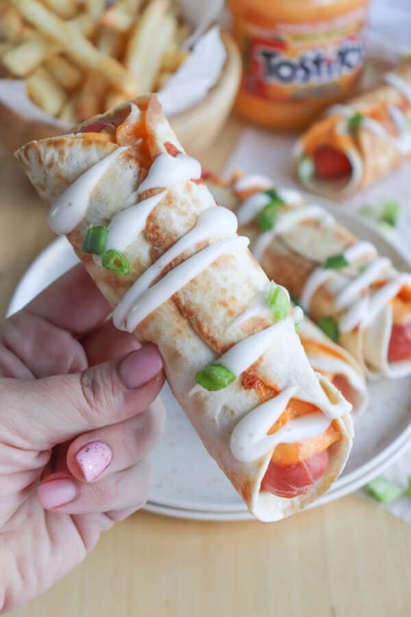 A hot dog topped with cheese sauce and wrapped in a tortilla, covered in crema and green onions being held in a hand.