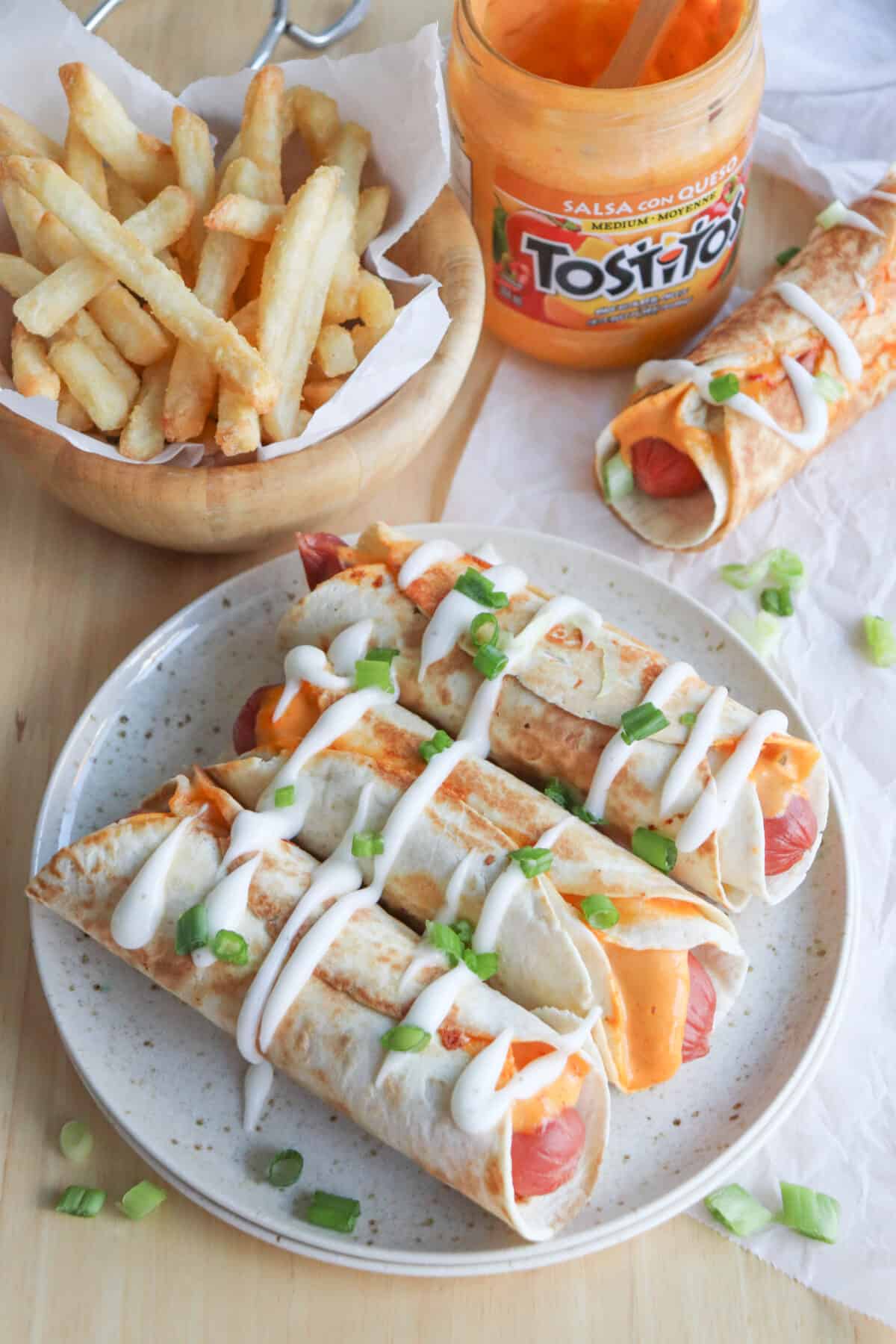 Three hot dogs wrapped in tortillas on a cream plate surrounded by a bowl of fries and a jar of open queso.