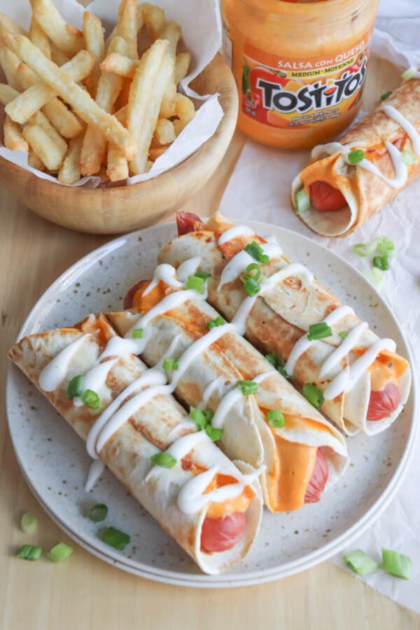Three hot dogs wrapped in tortillas on a cream plate garnished with crema and green onions.
