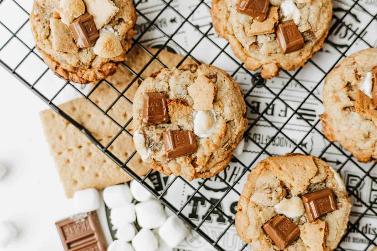 Cookies with chunks of chocolate, marshmallows, and graham cracker pieces are cooling on a black wire rack—The Best Smore Cookie Recipe—with extra chocolate, marshmallows, and graham crackers scattered nearby.