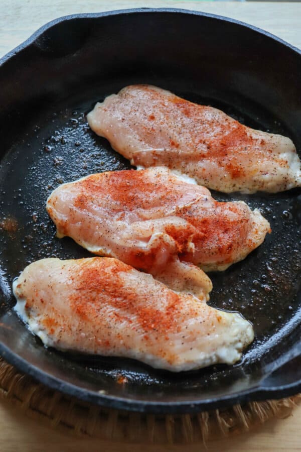 Three raw chicken breasts in a cast iron skillet with blackened seasoning.