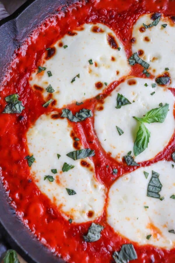 Close up of mozzarella cheese rounds in a bed of tomato sauce covering chicken in a cast iron skillet topped with fresh chopped basil.