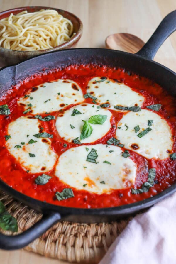 Mozzarella cheese rounds in a bed of tomato sauce covering chicken in a cast iron skillet topped with fresh chopped basil.