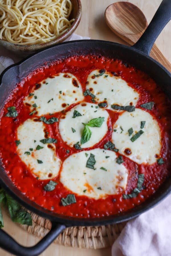 Mozzarella cheese rounds in a bed of tomato sauce covering chicken in a cast iron skillet topped with fresh chopped basil.