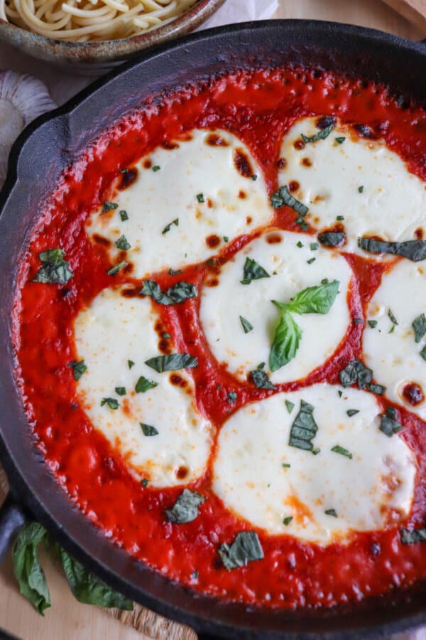 Close up of mozzarella cheese rounds in a bed of tomato sauce covering chicken in a cast iron skillet topped with fresh chopped basil.