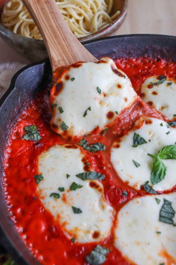 Wooden serving spoon scooping a mozzarella cheese round from a bed of tomato sauce covering chicken in a cast iron skillet topped with fresh chopped basil.