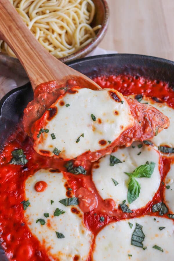Wooden serving spoon scooping a mozzarella cheese round from a bed of tomato sauce covering chicken in a cast iron skillet topped with fresh chopped basil.