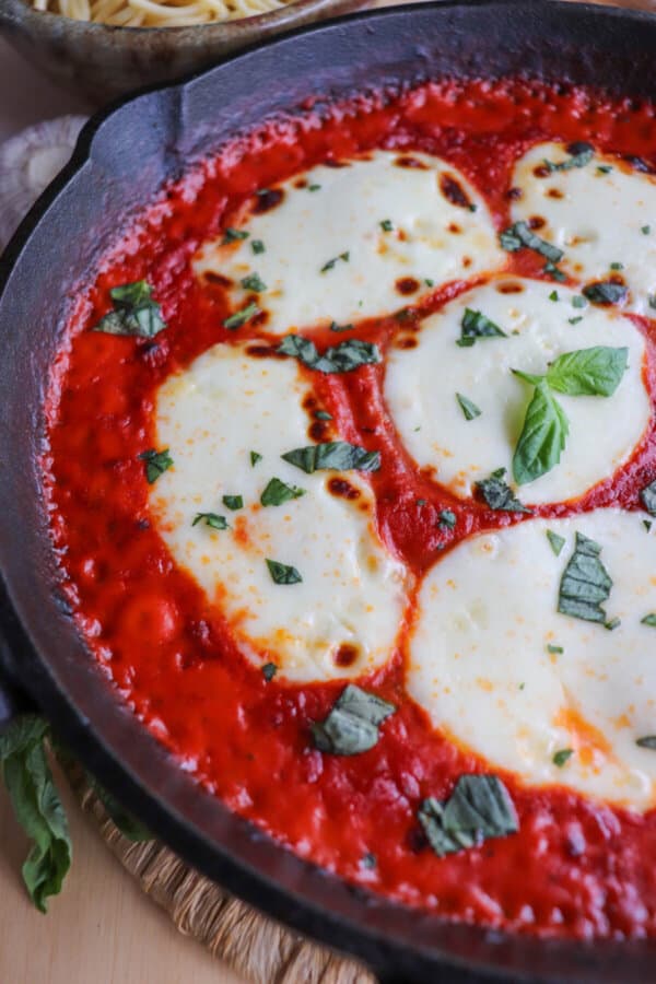 Close up of mozzarella cheese rounds in a bed of tomato sauce covering chicken in a cast iron skillet topped with fresh chopped basil.