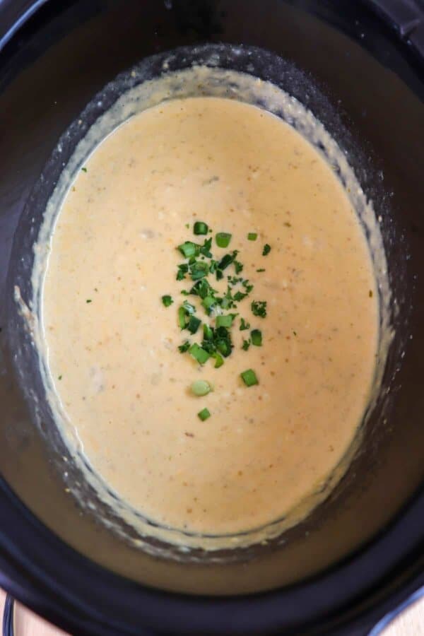 Creamy light orange cheese dip with diced jalapeno & green onion garnish in a black crockpot.