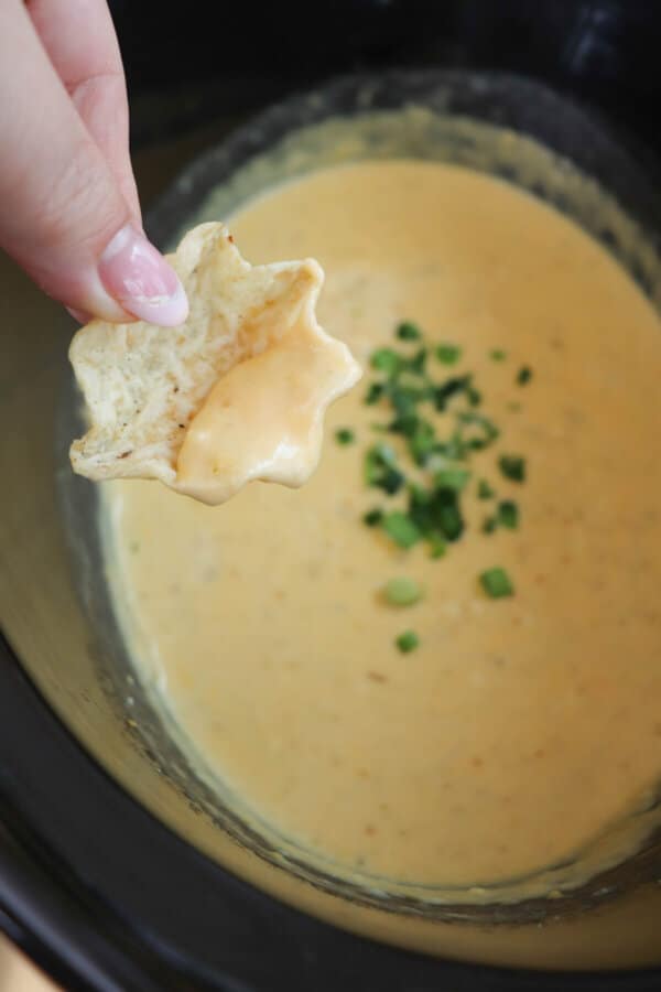Creamy light orange cheese dip with diced jalapeno garnish in a black crockpot with a tortilla chip being held in a hand.