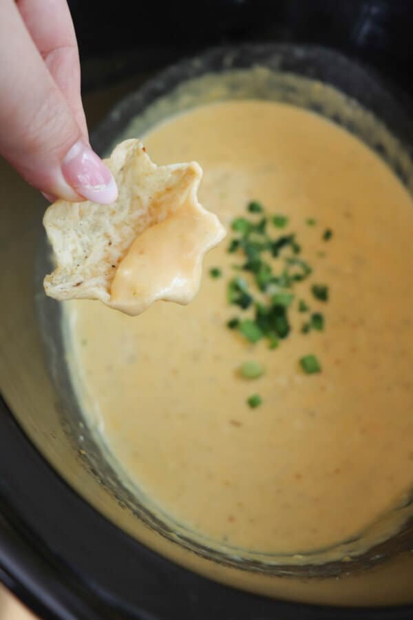 Creamy light orange cheese dip with diced jalapeno garnish in a black crockpot with a tortilla chip being held in a hand.