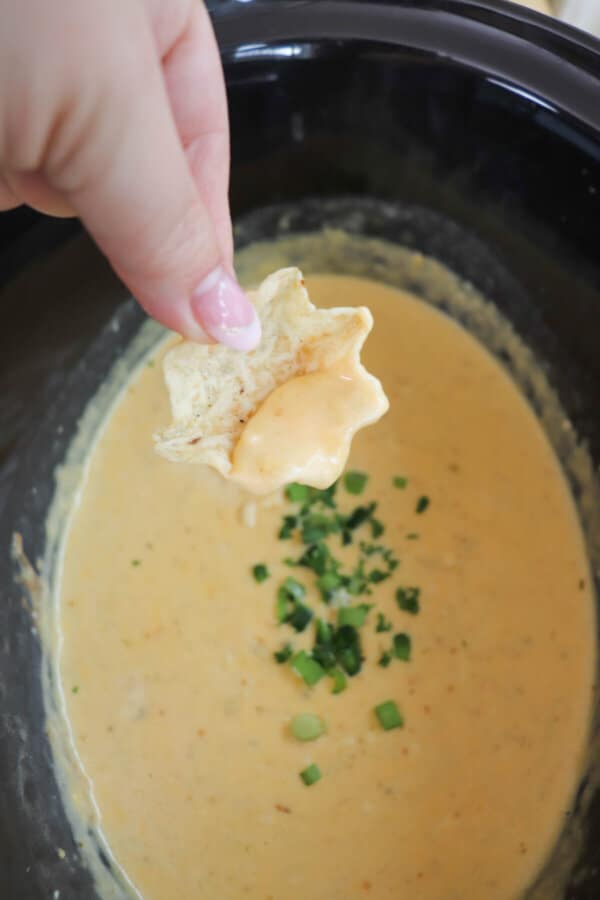 Creamy light orange cheese dip with diced jalapeno garnish in a black crockpot with a scoop on a tortilla chip in a hand.
