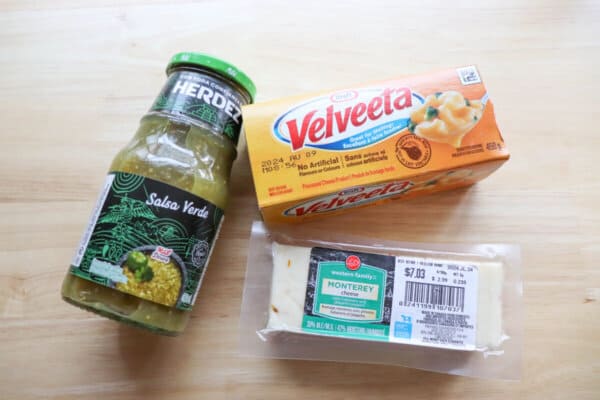 A jar of salsa verde, a package of Velveeta cheese and a block of pepper jack cheese on a wooden surface.