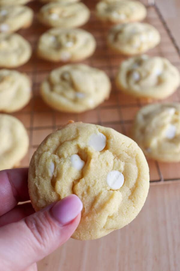 Yellow lemon cookie with white chocolate chips held in a hand with cookies on a wire rack in the background.