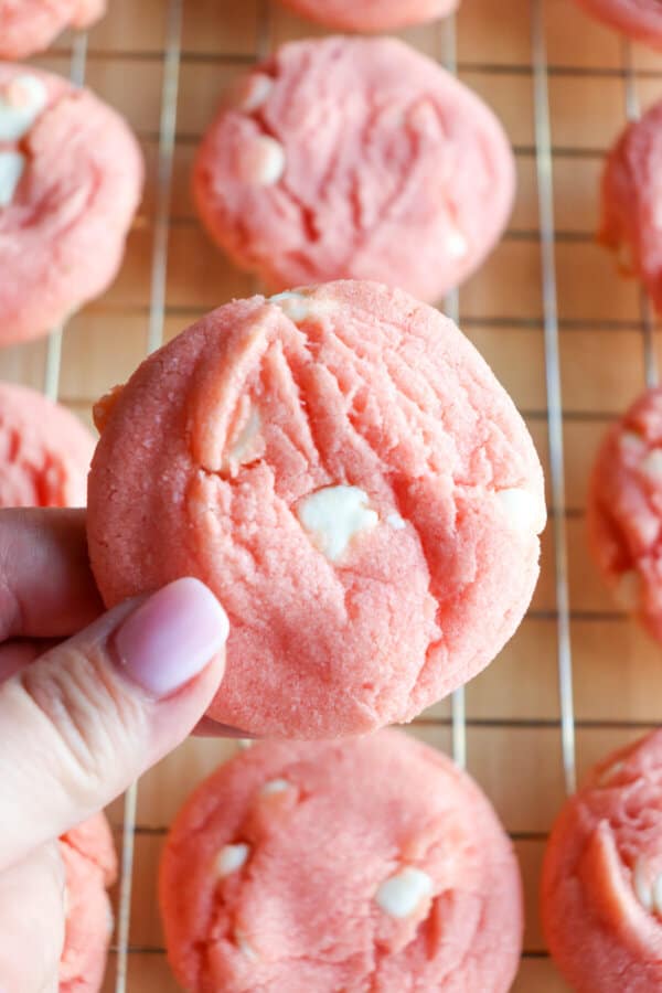 Pink cookie with white chocolate chips held in a hand with cookies on a wire rack in the background.