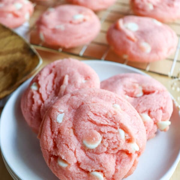 Pink cookies with white chocolate chips on a white plate with cookies on a wire rack in the background.