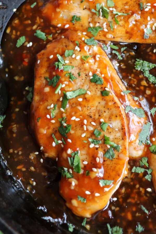 Close up of chicken breast covered in an orange sweet chili sauce topped with fresh cilantro and sesame seeds in a cast iron skillet.
