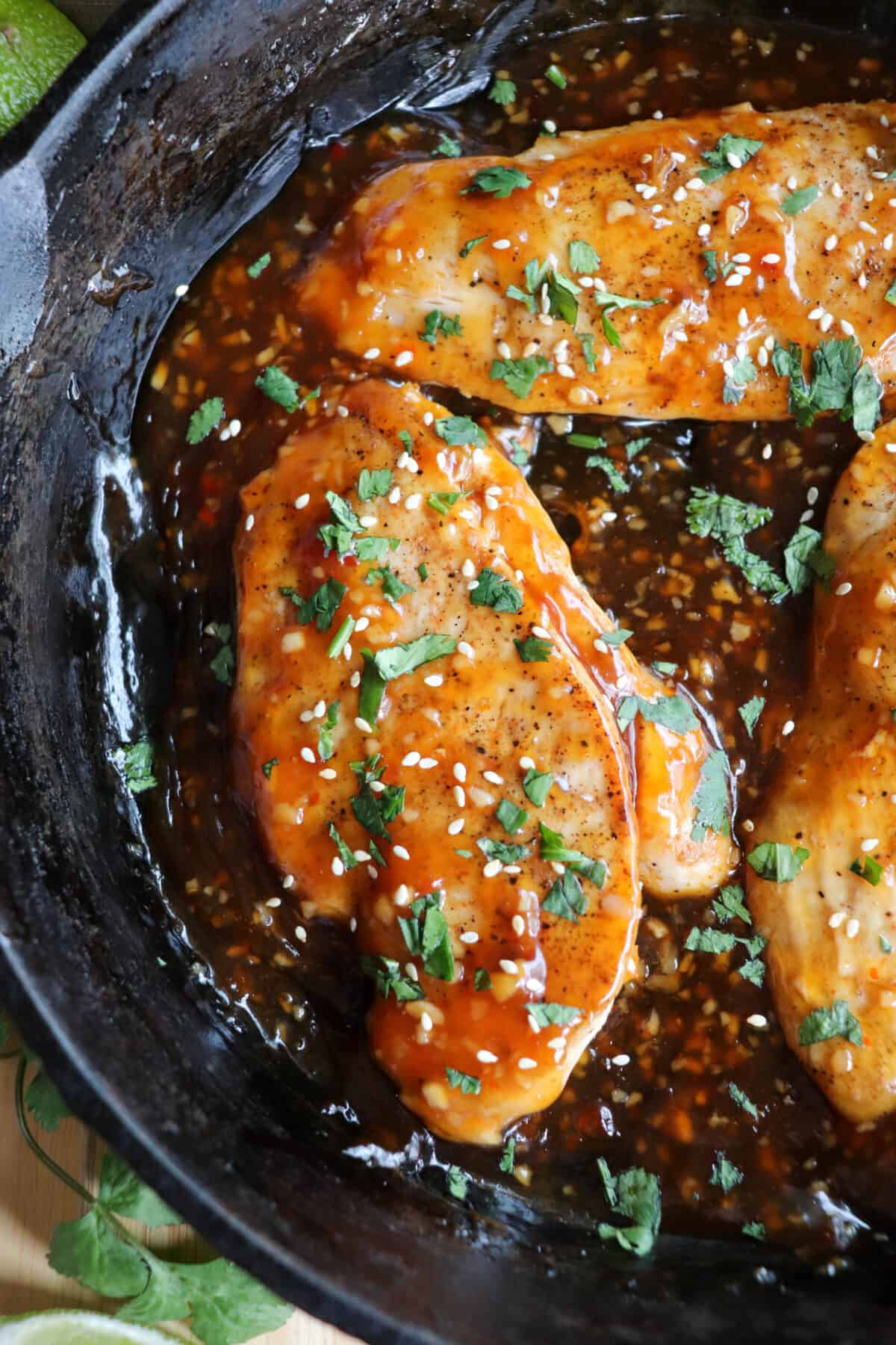 Close up of chicken breast covered in an orange sweet chili sauce topped with fresh cilantro and sesame seeds in a cast iron skillet.