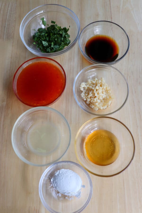 Ingredients to make sweet chili chicken in clear glass bowls on a wood backdrop.