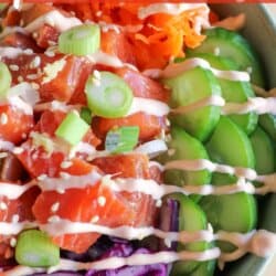 A green bowl with sliced cucumbers, grated carrot, sliced avocado, purple cabbage, diced red pepper with raw cubes of salmon topped with a drizzle of spicy mayo and diced green onion.