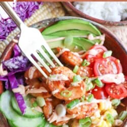 Cooked shrimp in a bowl surrounded by sliced cucumber, corn kernels, cherry tomatoes, sliced avocado and shredded red cabbage drizzled with spicy mayonnaise.