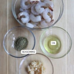 Ingredients for air fryer shrimp in clear glass bowls on a wooden table.