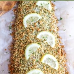 Salmon fillet covered in a crust of panko and herbs on a sheet of parchment paper topped with lemon slices for garnish.