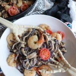 Purple and white linguine noodles in a cream sauce topped with shrimp and cherry tomatoes with skeleton hands in a white bowl.