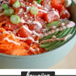 A green bowl with sliced cucumbers, grated carrot, sliced avocado, purple cabbage, diced red pepper with raw cubes of salmon topped with a drizzle of spicy mayo and diced green onion.