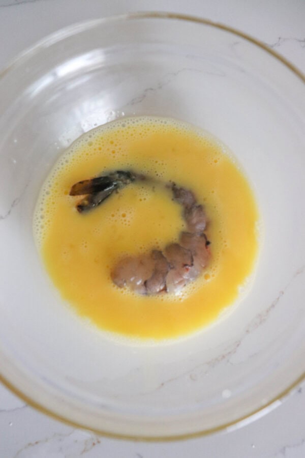 One raw jumbo shrimp in beaten egg mixture in a clear bowl.