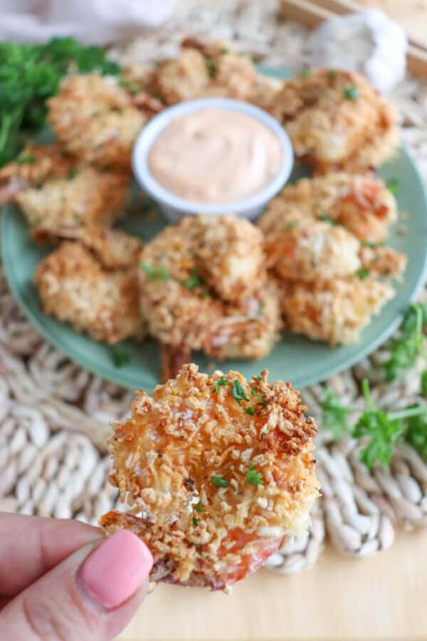 Panko shrimp held in hand with air fried panko shrimp on a green plate surrounding spicy mayo in the background.