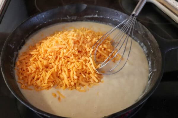 Cast iron skillet with creamy white rue and shredded cheddar cheese with a whisk in it.
