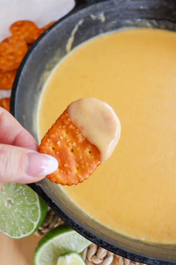 Fingers holding a pretzel cracker with cheese dip on it over a cast iron skillet of beer cheese sauce.