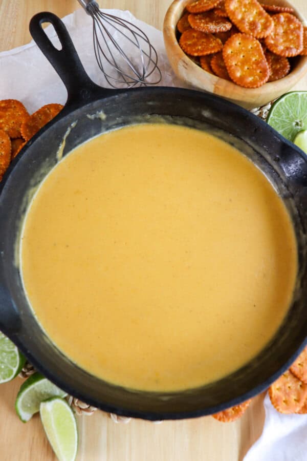 Black cast iron skillet with beer cheese sauce surrounded by pretzel crackers and fresh lime wedges on the table.