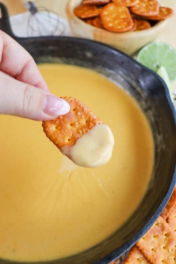 Fingers holding a pretzel cracker with cheese dip on it over a cast iron skillet of beer cheese sauce.