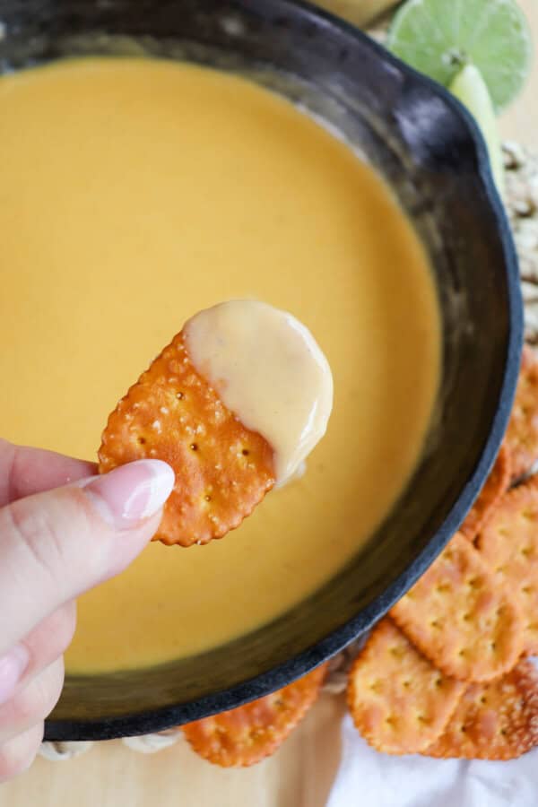 Fingers holding a pretzel cracker with cheese dip on it over a cast iron skillet of beer cheese sauce.