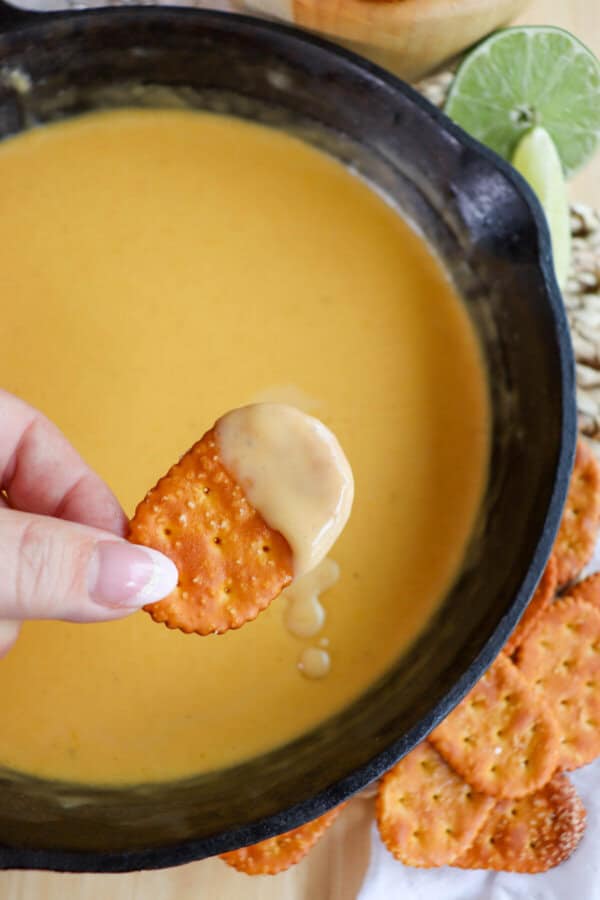 Fingers holding a pretzel cracker with cheese dip on it over a cast iron skillet of beer cheese sauce.
