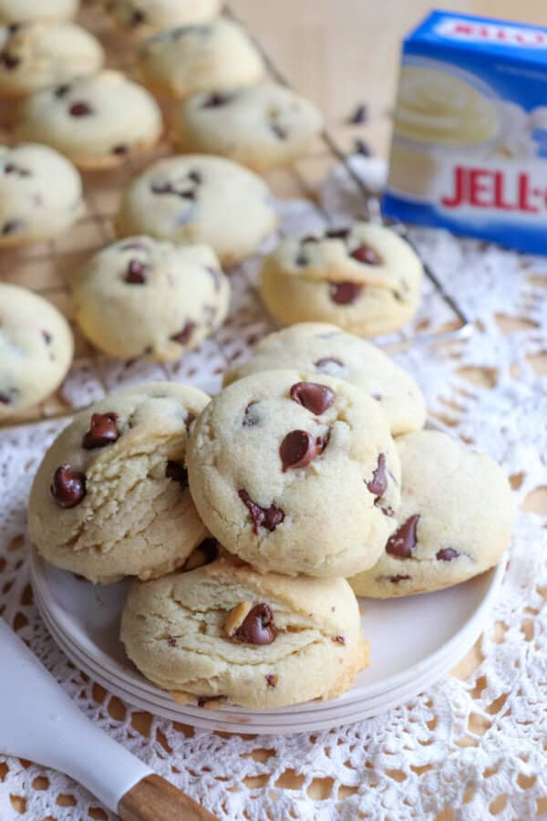 Chocolate chip cookies piled on a white plate with a wire rack of cookies in the background.