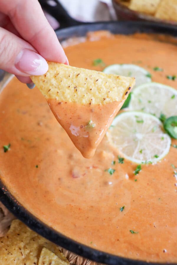 A triangle shaped tortilla chip being dipped into a skillet of cheesy queso with slices of lime on top in the background.