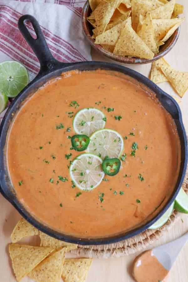 Cheesy dip in a cast iron skillet with three slices of lime, two round slices of jalepeno and fresh parsley for garnish in the center.  The skillet is surrounded by fresh lime wedges and tortilla chips on a wooden table.