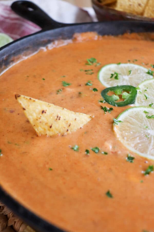 A triangle shaped tortilla chip being dipped into a skillet of cheesy queso with slices of lime on top in the background.
