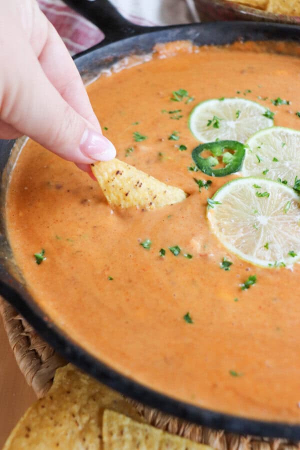 A triangle shaped tortilla chip being dipped into a skillet of cheesy queso with slices of lime on top in the background.