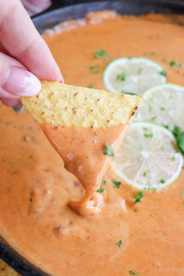 A triangle shaped tortilla chip being dipped into a skillet of cheesy queso with slices of lime on top in the background.